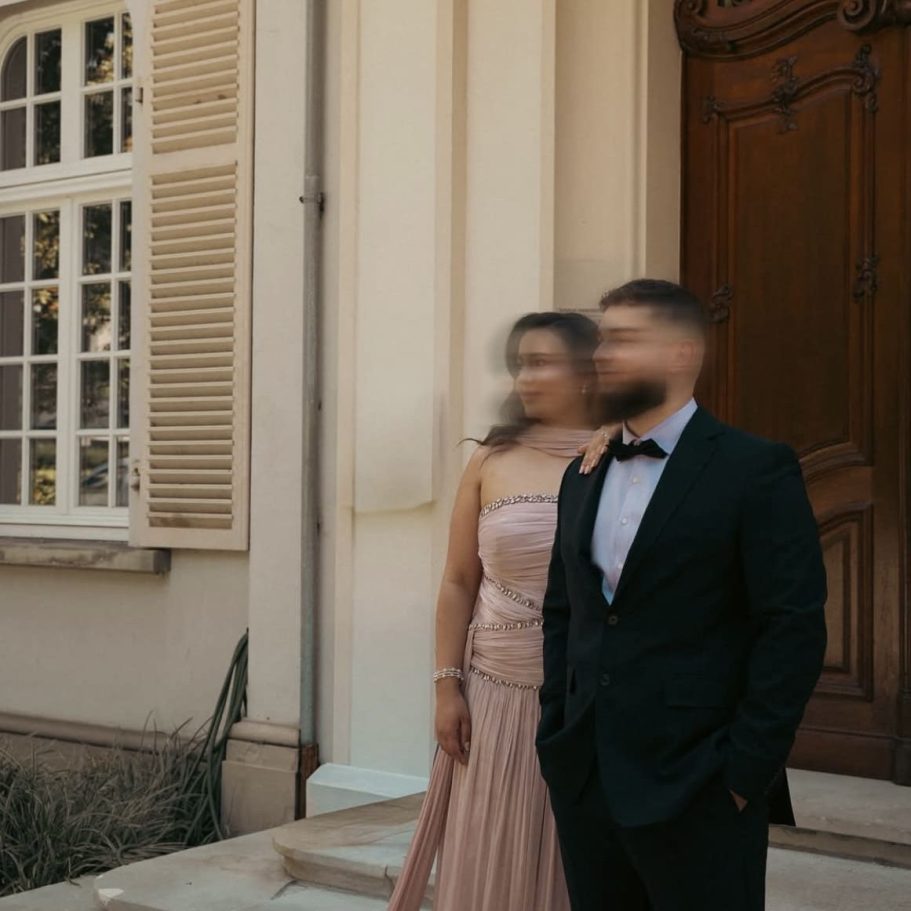 Ein Mann im Smoking und eine Frau im Abendkleid stehen vor einem Haus, leicht unscharf.